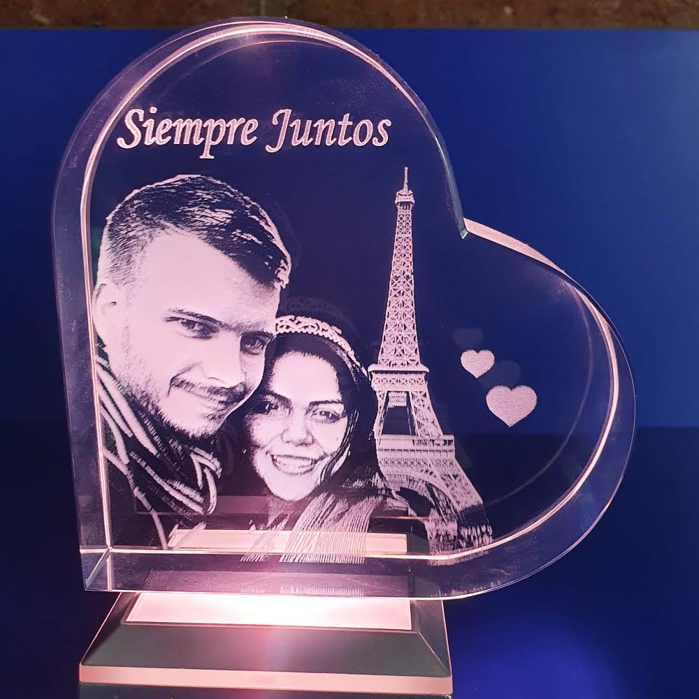 Un bloque de cristal en forma de corazón grabado con láser. En el interior hay un grabado en 3D de una pareja sonriente, la Torre Eiffel y dos corazones flotantes. El texto "Siempre Juntos" está grabado sobre ellos. El cristal está iluminado desde abajo por una base metálica que brilla en color rosa/púrpura, y el fondo es de color azul marino intenso